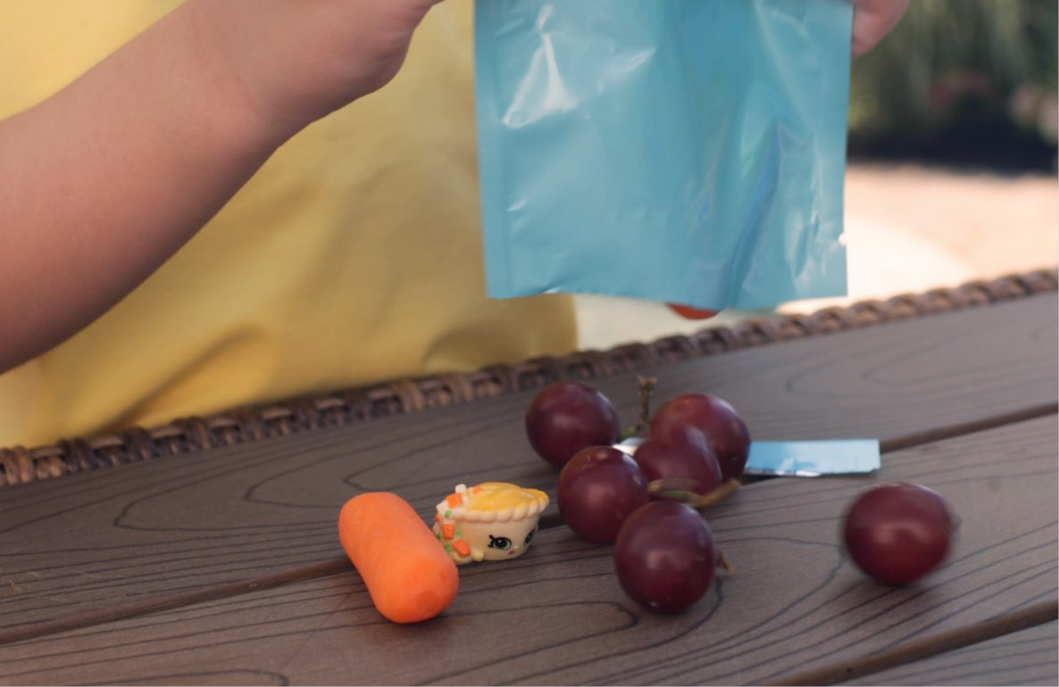 Child's hand holding blue paper bag above wooden surface with small toy figures and dark grapes during snack bag surprise activity