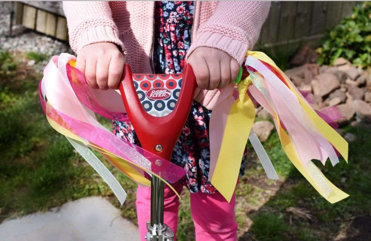 Child modeled by a child holding red handlebar streamers with pink and yellow ribbons attached, on a bicycle outdoors