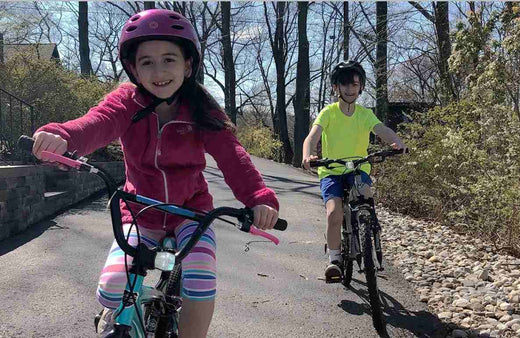 Two children riding bikes on a paved path outdoors; child on left wears burgundy jacket and helmet, child on right wears neon yellow shirt and helmet