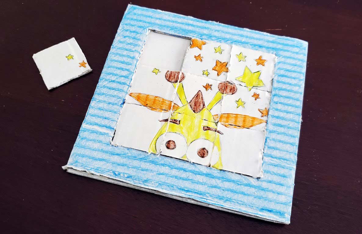 Handmade cardboard slide puzzle with blue striped border featuring a yellow creature with orange wings and brown details surrounded by colorful stars