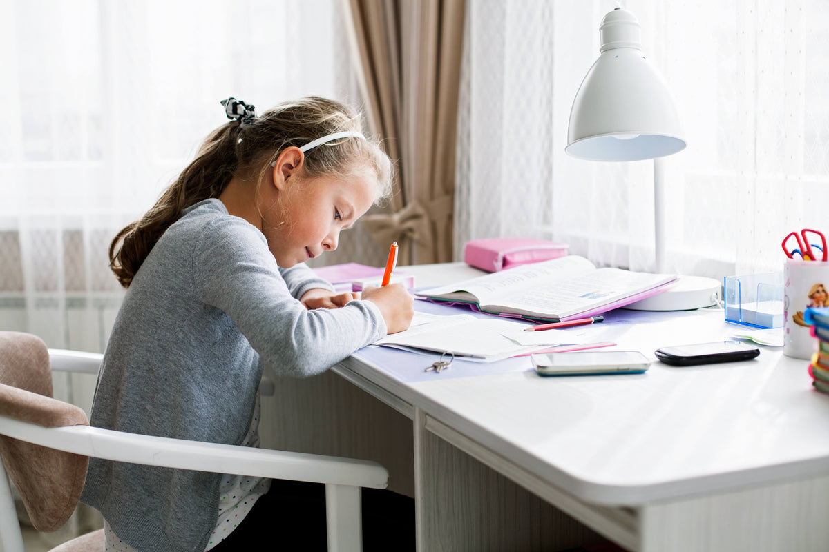 Child modeled by a child sitting at white desk, writing in notebook with pencil under white pendant lamp, with school supplies and open books nearby