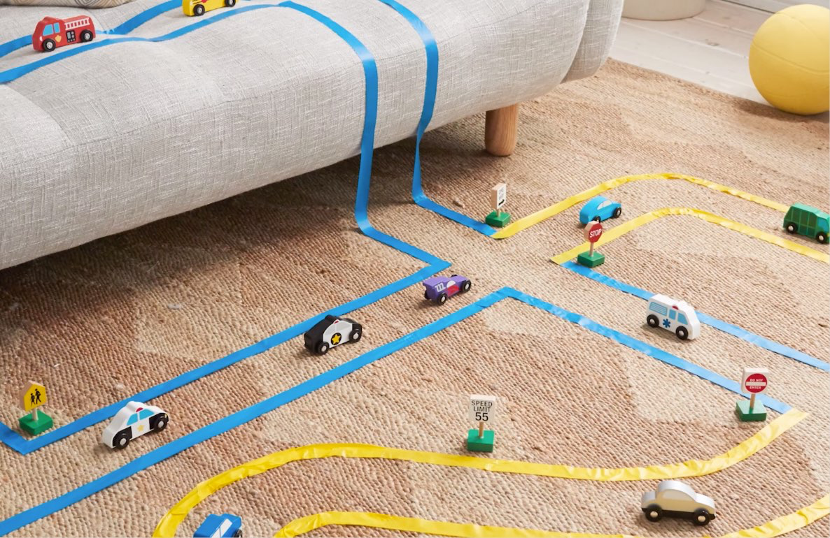 Child's DIY race track made with blue and yellow masking tape on a beige rug, featuring toy cars, traffic signs, and a speed limit sign, with a gray sofa and ye