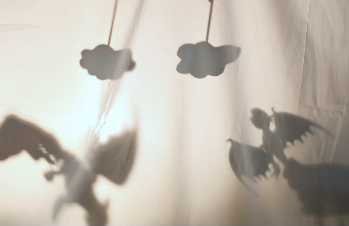 Silhouettes of winged toys and cloud shapes cast against a white fabric backdrop in a shadow theater setup