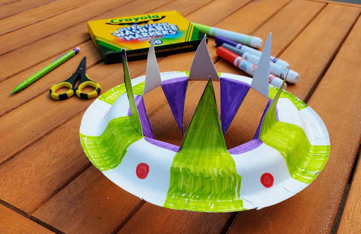 DIY flying saucer craft made from paper plates colored with markers, featuring green fringe, purple pointed spires, and red details, surrounded by Crayola marke
