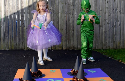 Two children on a wooden fence background playing DIY Halloween ring toss game with orange and purple cards, cone towers, and Halloween decorations on black mat