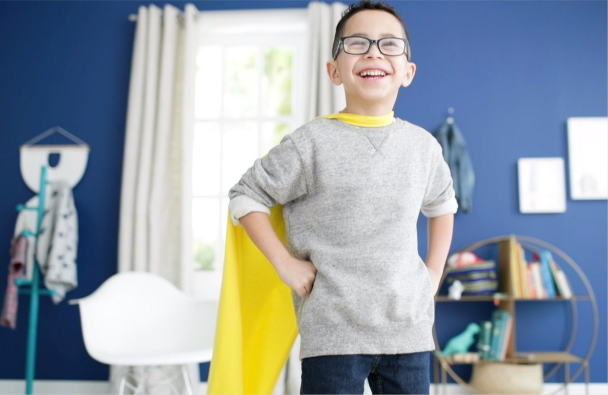 Child wearing glasses and a gray sweatshirt with a yellow cape, smiling in a blue bedroom with bookshelves and toys