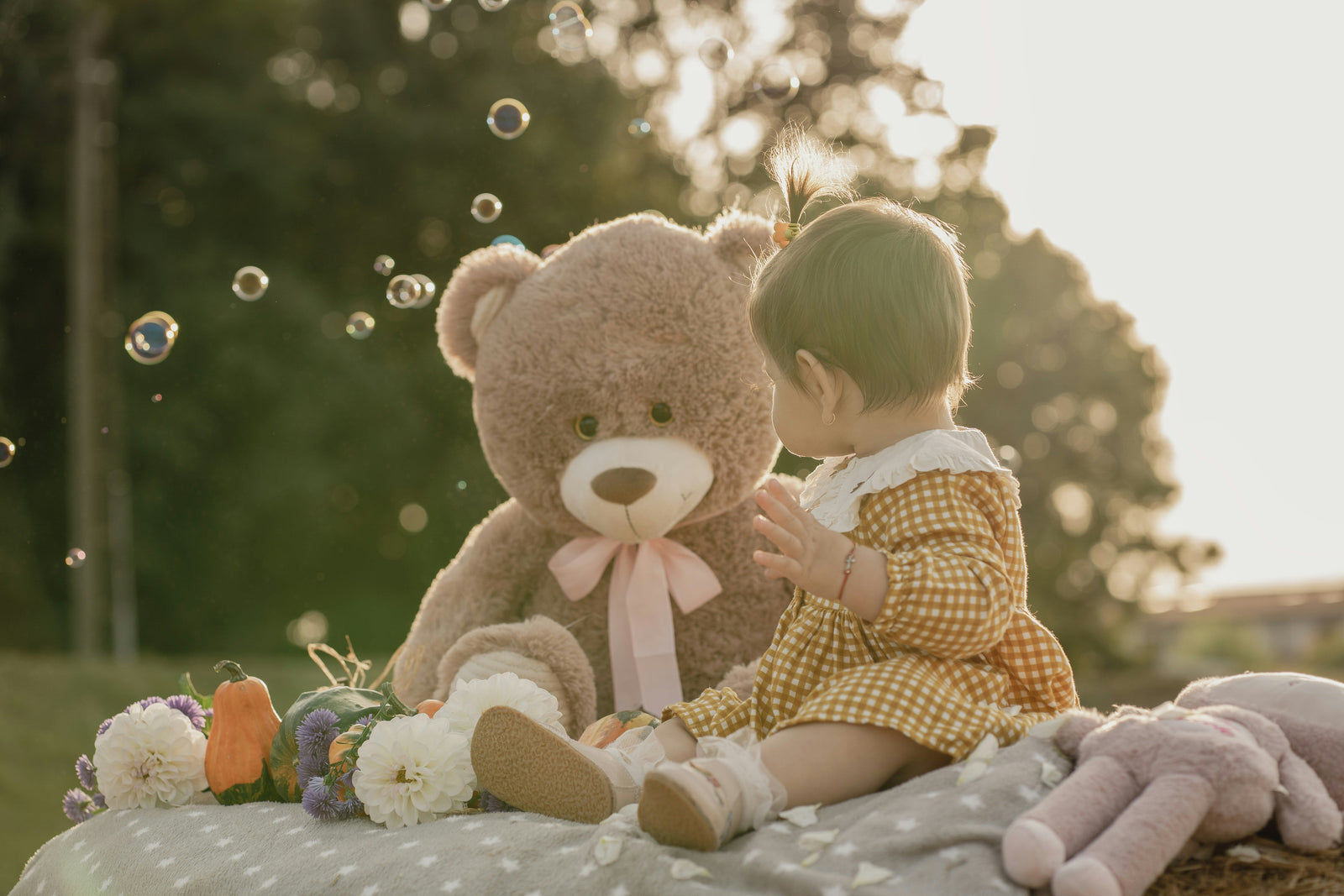 Toddler modeled by a child in yellow gingham dress sitting beside a soft gray teddy bear with pink ribbon, surrounded by white flowers and bubbles in a sunlit g