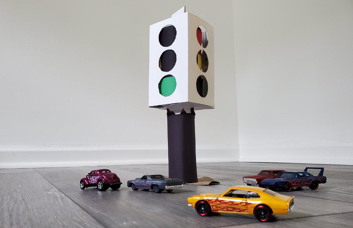 DIY cardboard traffic light with red, yellow, and green lights on a black pole, surrounded by toy cars on a floor