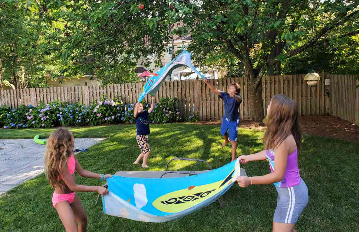 Children playing with blue and yellow kites in a backyard on grass, with wooden fence and flowering bushes in background