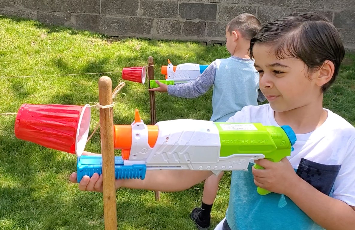 Children playing with water blasters in a backyard, aiming at targets mounted on a wooden pole in the grass