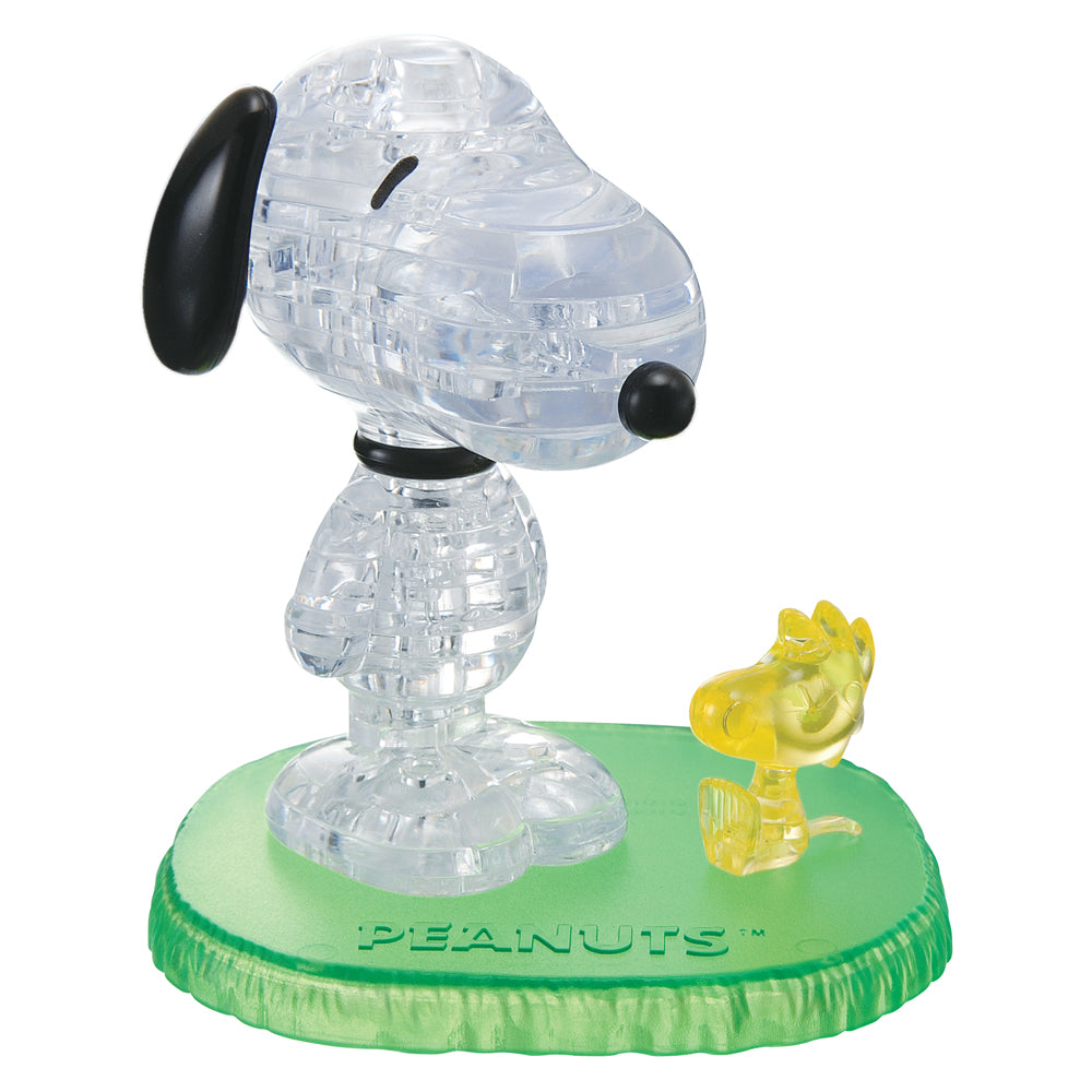 Clear crystal 3D puzzle of Snoopy with black ears and nose, standing on green base with yellow Woodstock figure, labeled PEANUTS