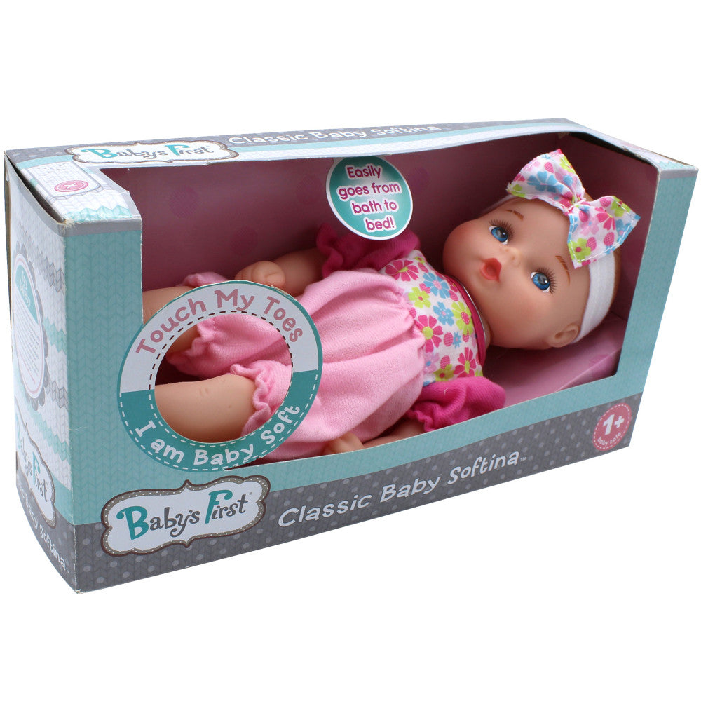 Baby's First Classic Softina doll in packaging, soft-bodied baby doll with blue eyes and colorful bow, wearing pink outfit, displayed in turquoise and burgundy