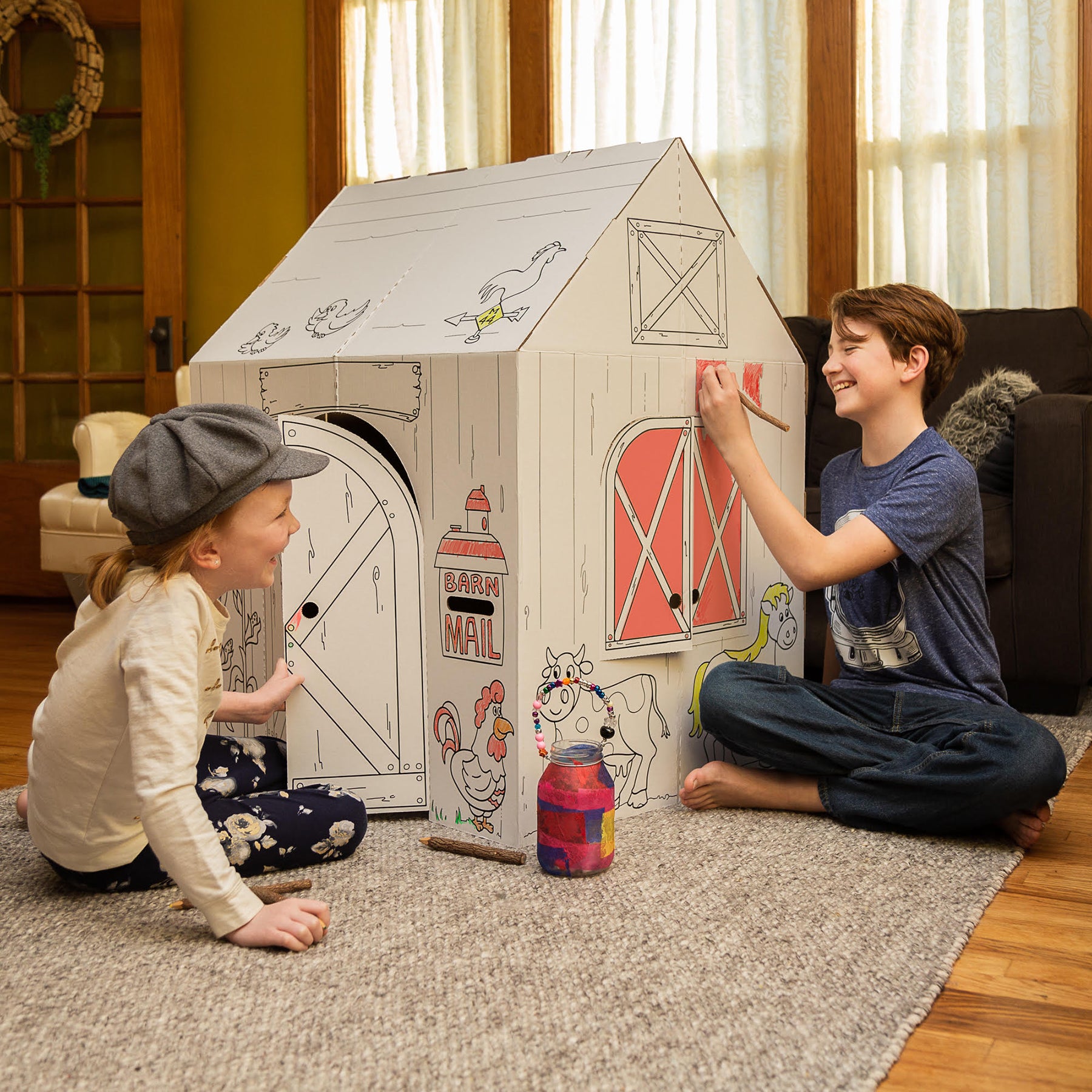Two children coloring a cardboard barn playhouse together on the floor, modeled by children in a home setting