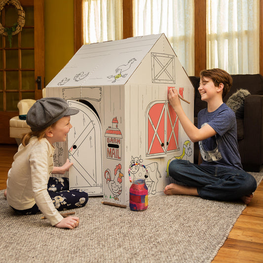 Two children coloring a cardboard barn playhouse together on the floor, modeled by children in a home setting
