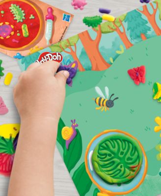 Child's hand holding Play-Doh Light and Look Microscope Playset with colorful nature-themed playmat featuring flowers...