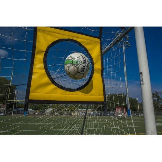 Soccer ball passing through yellow target with black circle opening attached to blue goal net on outdoor field