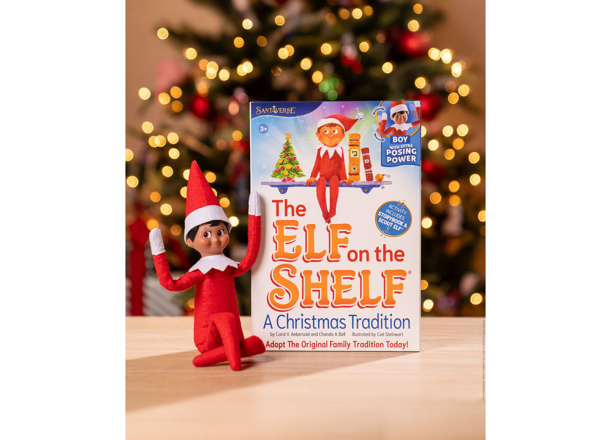 Elf on the Shelf Scout Elf Boy figure in red suit next to storybook, Christmas tree and lights in background