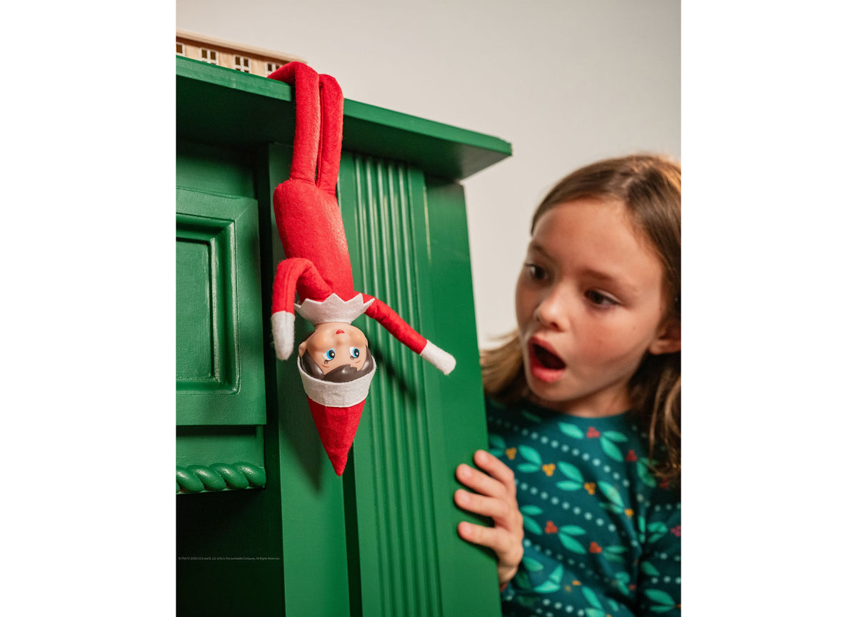 Scout Elf with blue eyes in red and white outfit perched on green shelf, child reacting with surprise