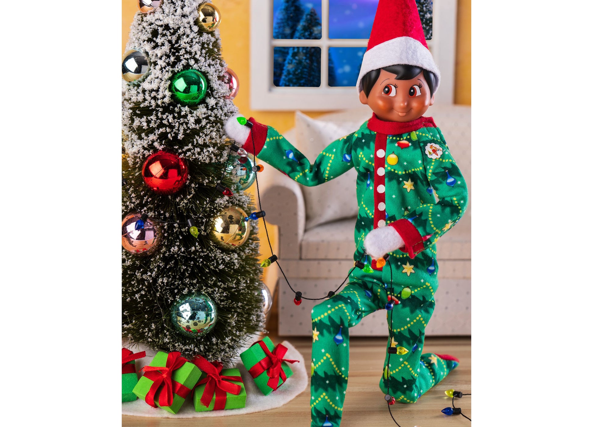 Elf on the Shelf in green Christmas pajamas with red trim, posing by decorated tree