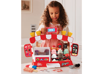 Girl holding open sign above red and white Casdon Ice Cream Parlor playset with freezer, ice cream flavors, and acces...