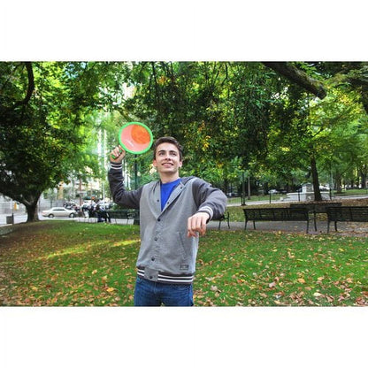 Child modeled by a boy in a park throwing a green and orange Zingo Dual Bat and Ball Set disc, with trees and benches in background