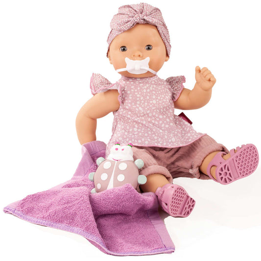 Baby doll with pink polka dot dress, turban headwrap, and pacifier, sitting on purple blanket with small plush ladybug toy and pink bath accessories