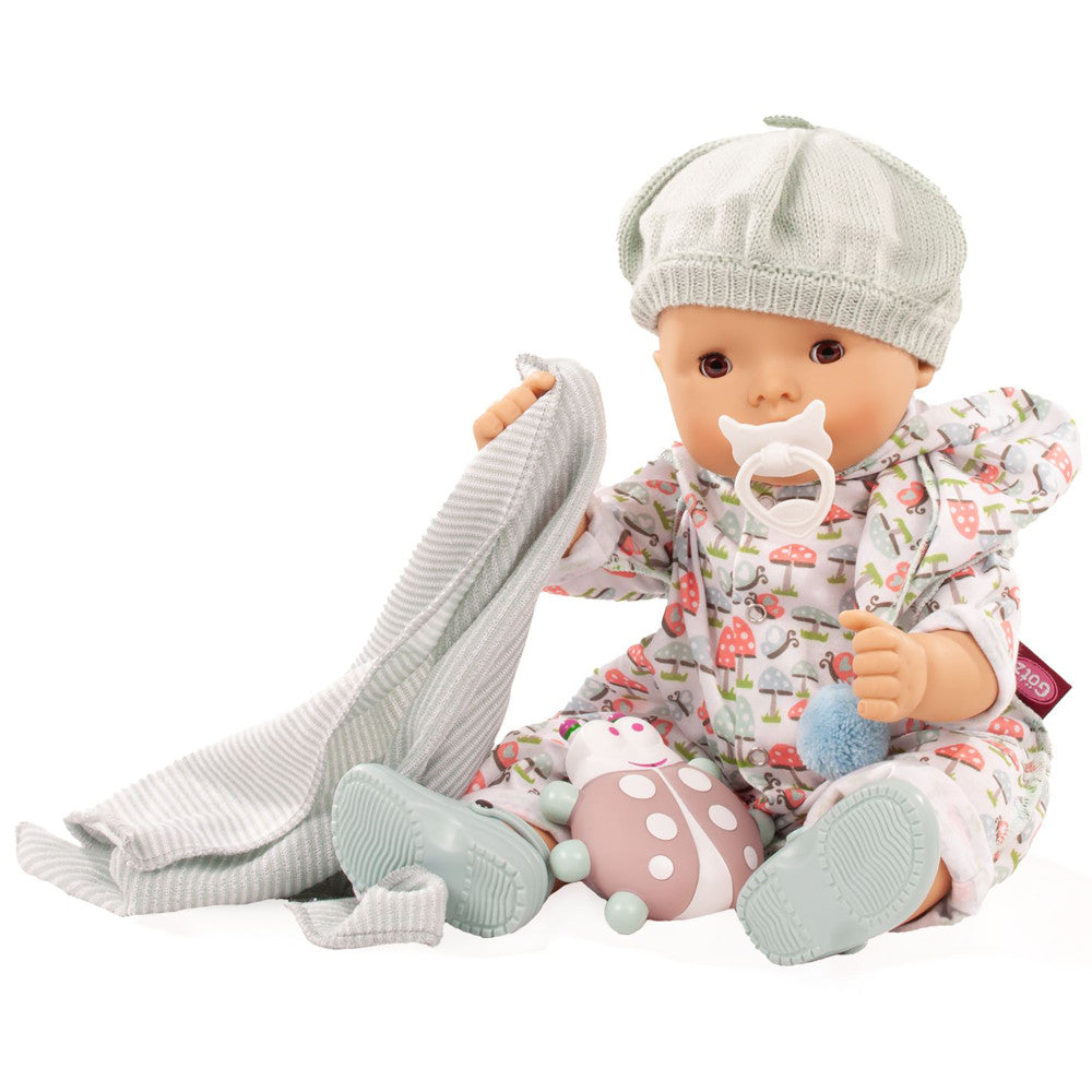 Baby doll wearing white knit hat and mushroom-print outfit with white pacifier, holding gray knit blanket and pink mushroom rattle toy
