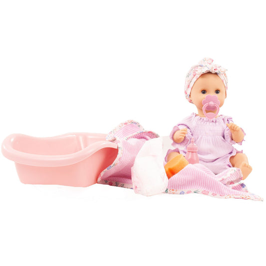 Baby doll in pink dress and bonnet with pacifier, seated next to pink bath tub and folded washcloth with accessories