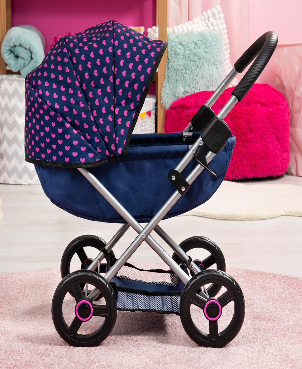 Doll pram stroller with navy blue frame, pink heart-patterned canopy, black wheels with pink accents, displayed in styled room setting with plush pillows and bl