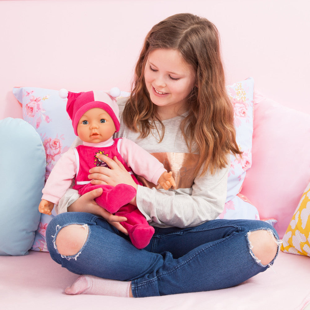 Child modeled by a girl holding a baby doll in hot pink outfit and matching hat, seated on pink pillows against a pink background