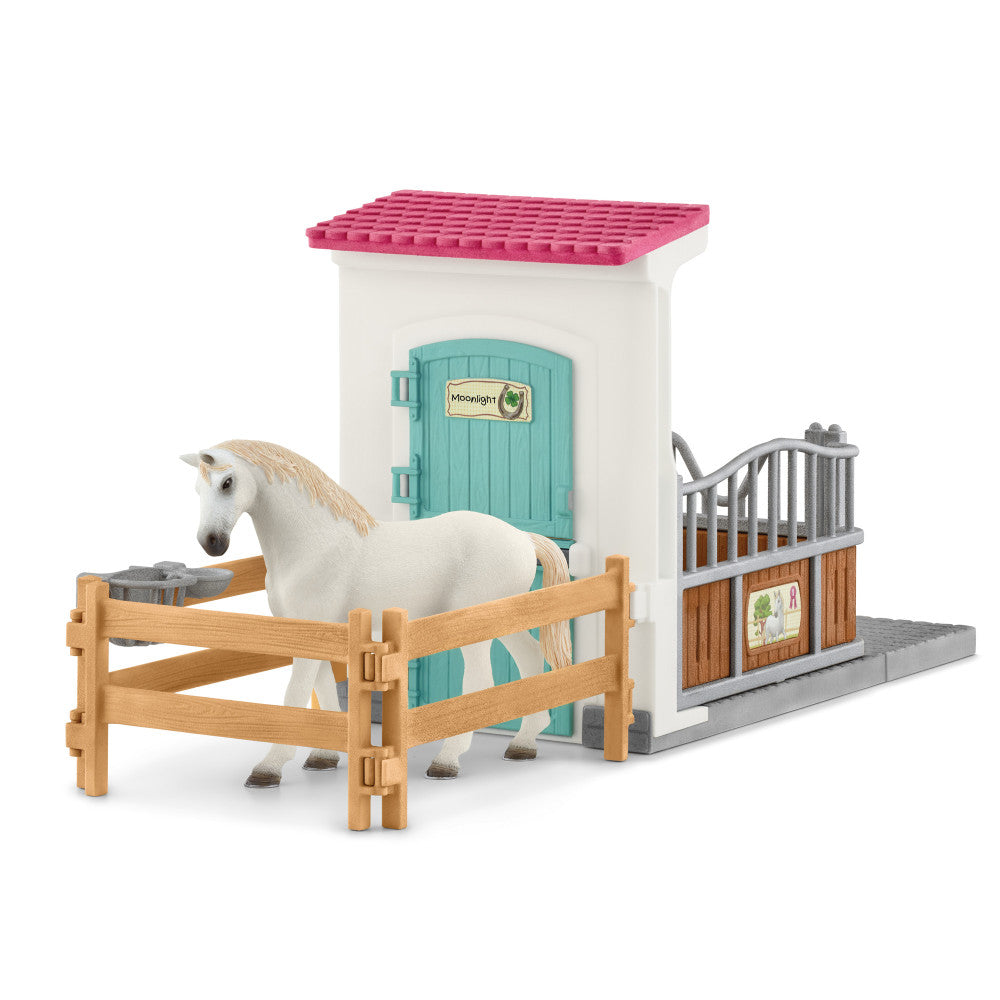White horse with tan mane standing in wooden fence stall beside pink-roofed white barn building with turquoise door and gray metal feed gate