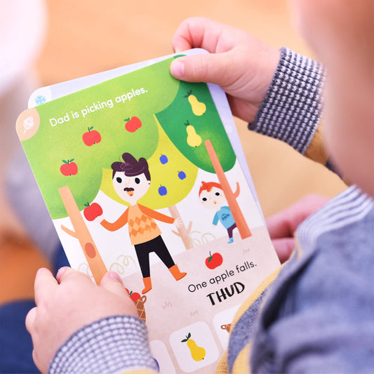 Child's hands holding open a colorful picture book with text 'Dad is picking apples. One apple falls. THUD.' showing illustrated father and child under an apple
