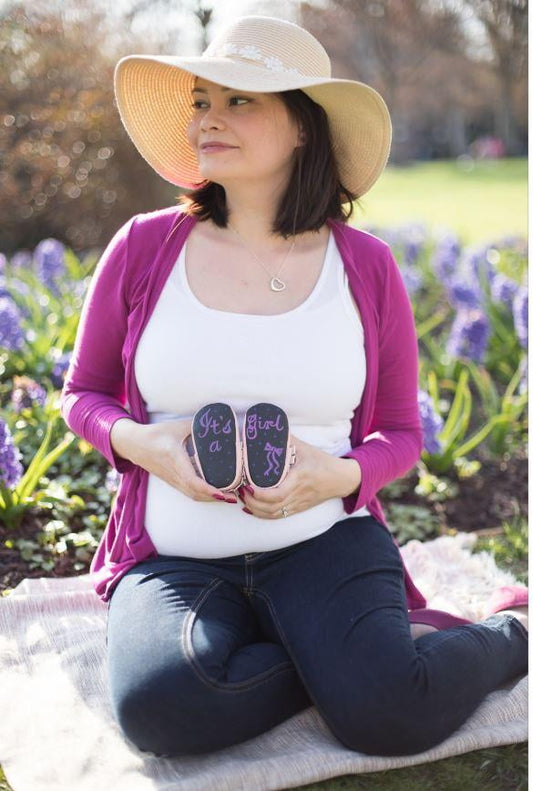 Pregnant woman on model wearing pink cardigan and white hat, holding chalkboard sole moccasins in garden setting with purple flowers