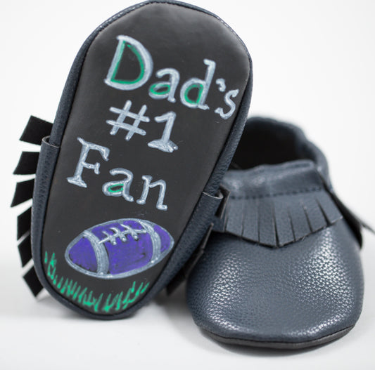 Black moccasin slippers with fringe trim and rubber sole printed with 'Dad's #1 Fan' text and football graphic