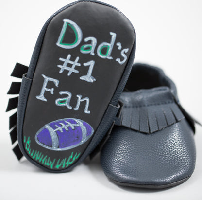 Gray moccasin slipper with black fringe trim and chalkboard sole featuring 'Dad's #1 Fan' text and football illustration, shown from sole and side view