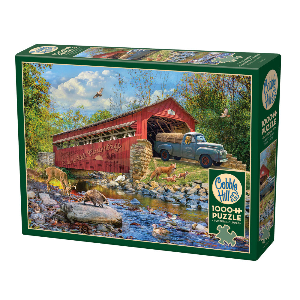 Cobble Hill 1000+ piece jigsaw puzzle box featuring rural scene with red covered bridge, vintage truck loaded with hay, deer and wildlife by stream