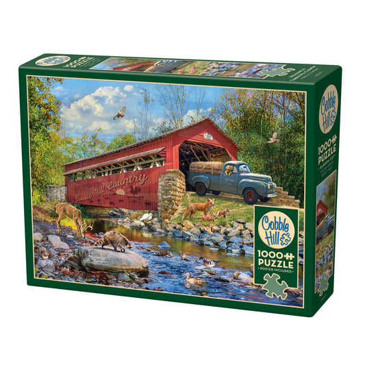 Cobble Hill 1000+ piece jigsaw puzzle box featuring rural scene with red covered bridge, vintage truck loaded with hay, deer and wildlife by stream