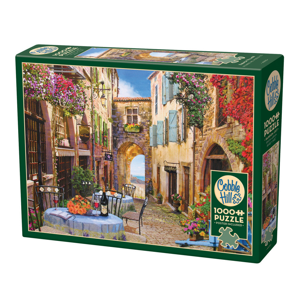 Cobble Hill 1000-piece puzzle box featuring a European village street scene with colorful flowering vines, stone buildings, outdoor café table, and ocean view