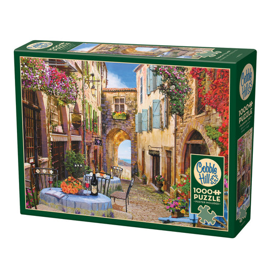 Cobble Hill 1000-piece puzzle box featuring a European village street scene with colorful flowering vines, stone buildings, outdoor café table, and ocean view
