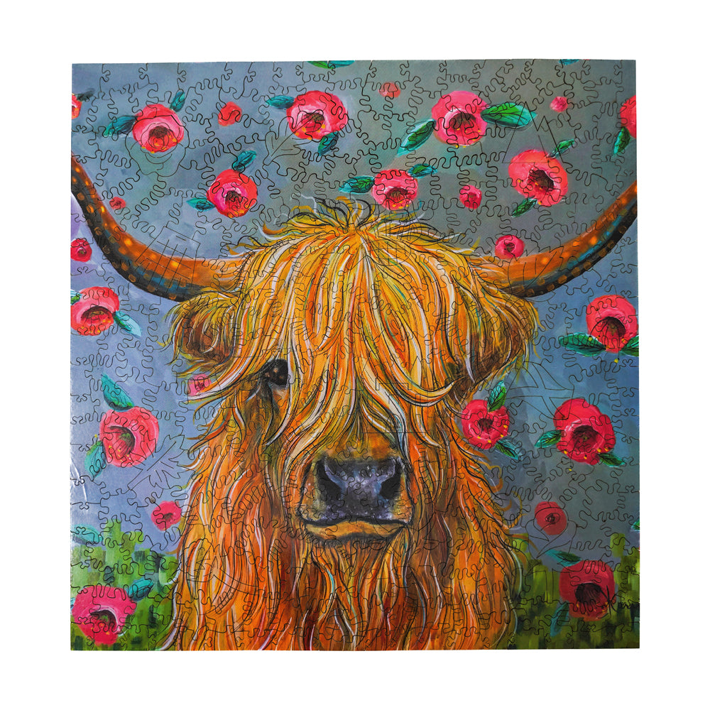 Wooden jigsaw puzzle featuring an illustrated Highland cow with long orange hair and horns surrounded by pink flowers and green foliage on a blue puzzle-piece b
