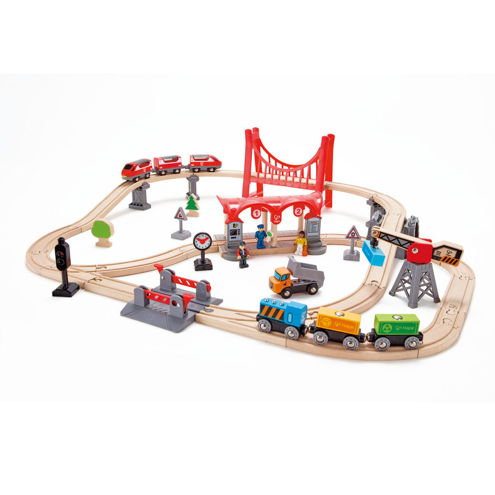Wooden train set with curved beige tracks, red bridge, red train cars, cargo trucks, traffic signs, buildings, and figurines on plain background