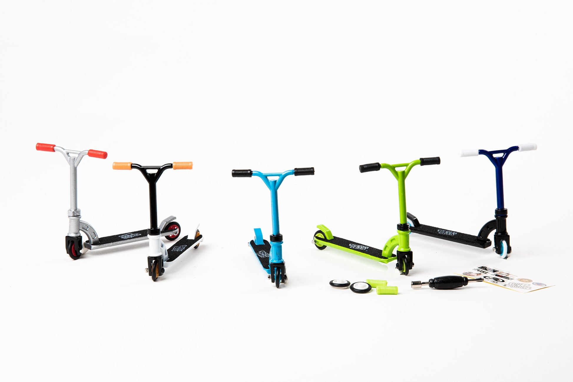 Five colorful finger scooters in silver, black, black and orange, blue, and lime green with black accents, displayed on white background with accessories includ
