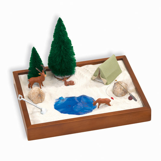 Wooden sandbox with white sand containing two green evergreen trees, brown deer figurine, green canvas tent, rocks, blue water feature, and camping accessories