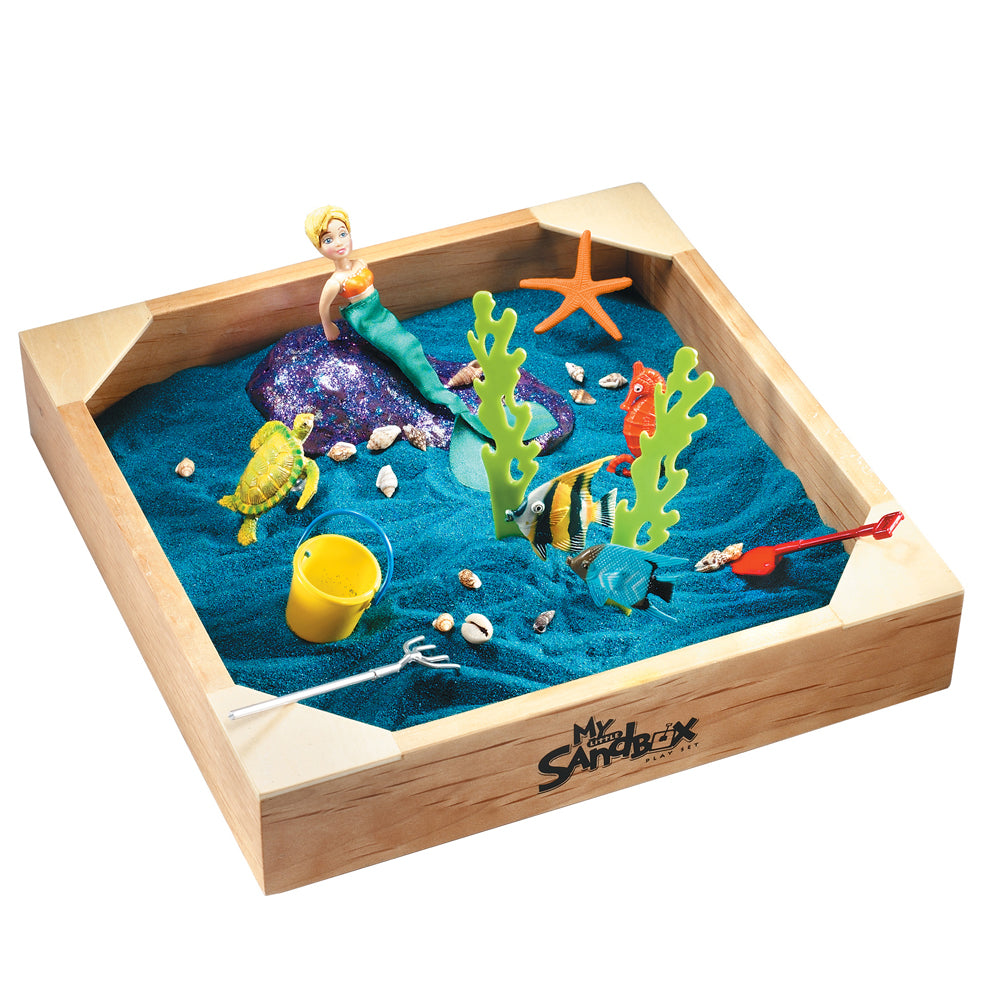 Wooden sandbox with blue sand containing mermaid figurine, sea turtle, tropical fish, starfish, coral, shells, yellow bucket, and fishing net toy