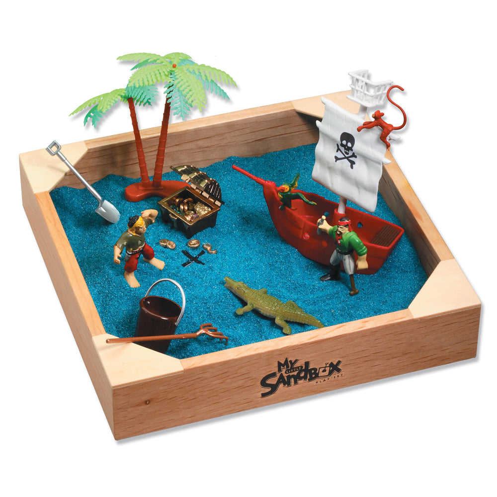 Wooden sandbox with blue sand containing pirate-themed figurines, red ship, palm tree, treasure chest, skull flag, crocodile, shovel, and bucket
