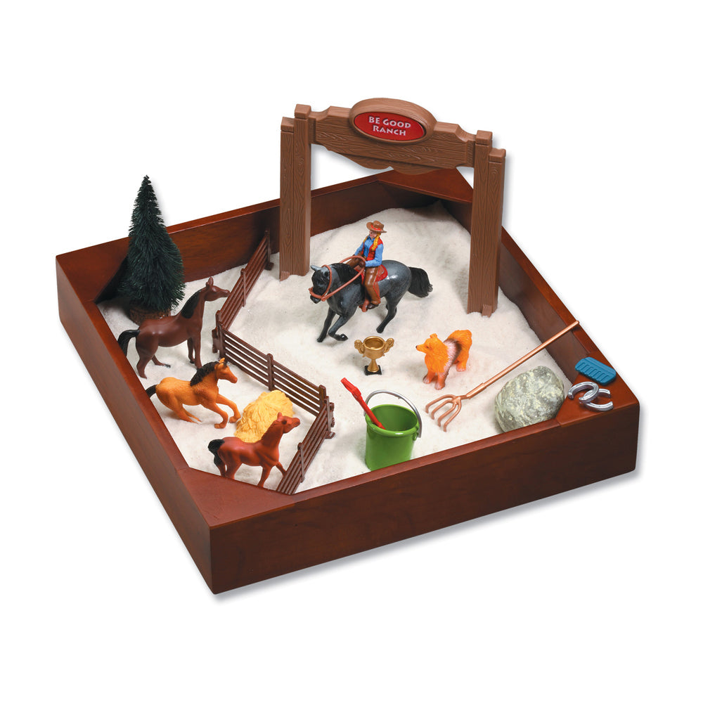 Brown wooden sandbox play set with white sand, cowboy figurine on black horse, multiple horse figures, ranch gate with 'Be Good Ranch' sign, fencing, tree, and