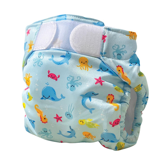 Blue swim nappy with colorful sea creatures including whales, jellyfish, starfish, seahorses, and fish, with white waterproof panels and blue trim