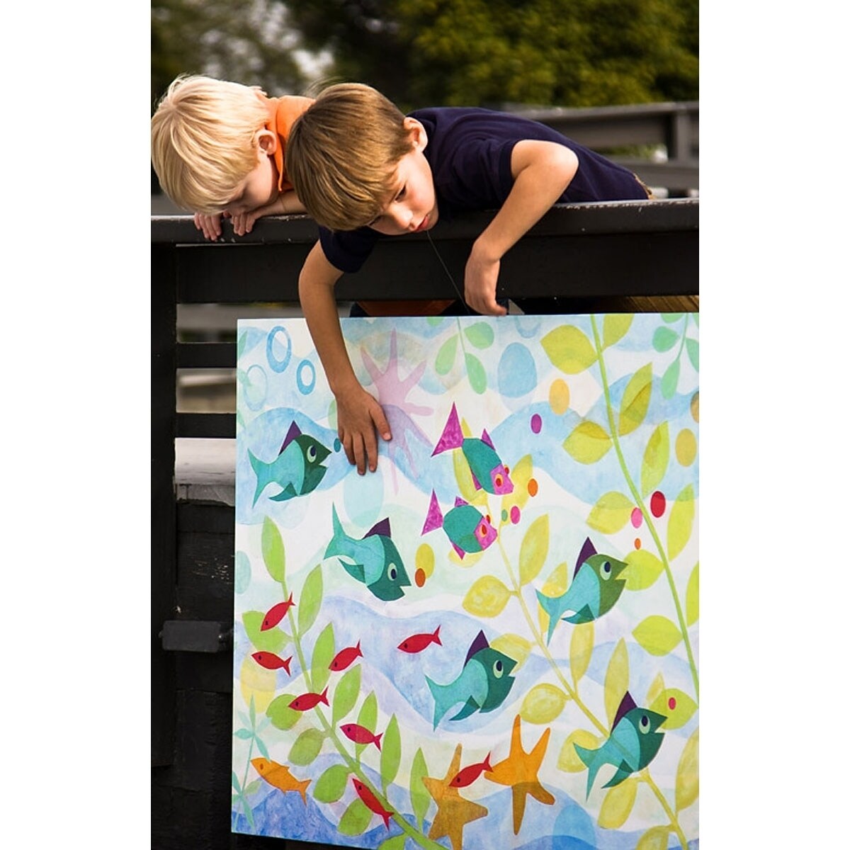 Two children leaning over a colorful canvas displaying friendly fish, aquatic plants, and starfish in turquoise, green, yellow, and red tones