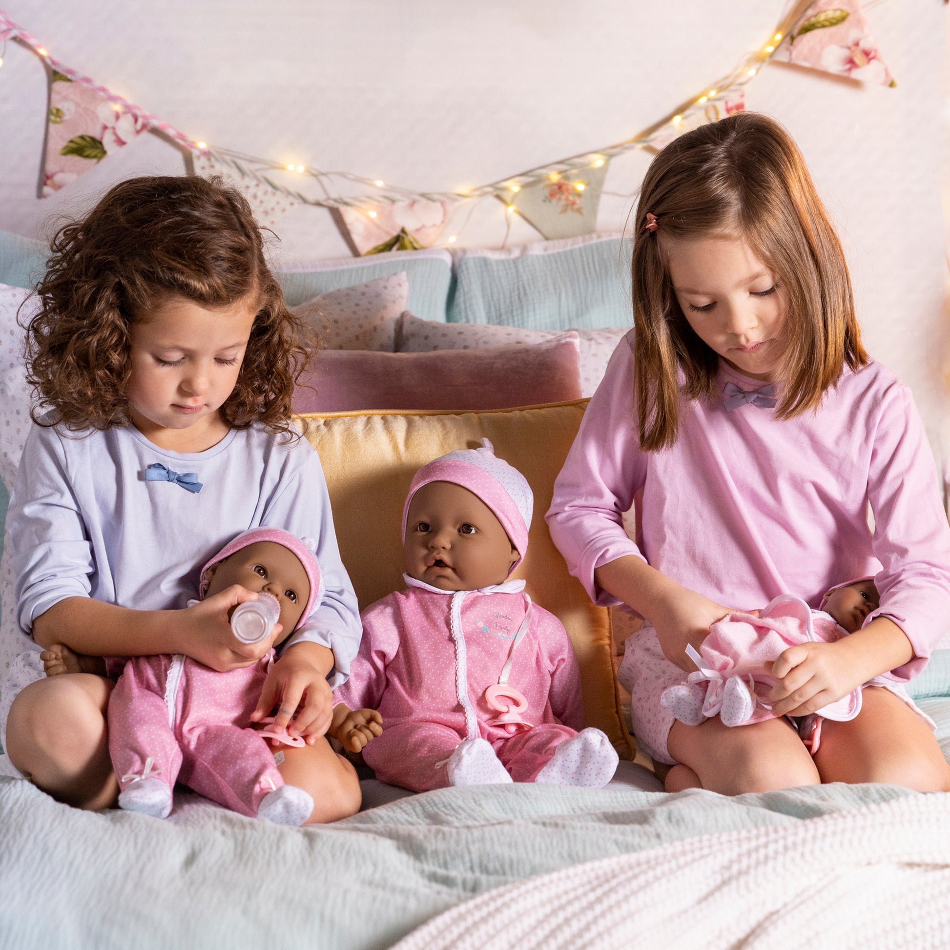 JC Toys La Baby Hispanic doll in pink outfit with pacifier, held by two girls on bed