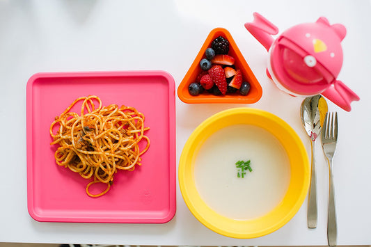 Colorful kid-friendly dining set with pink square plate holding pretzel snacks, yellow bowl with soup and green garnish, orange triangle bowl with berries, pink